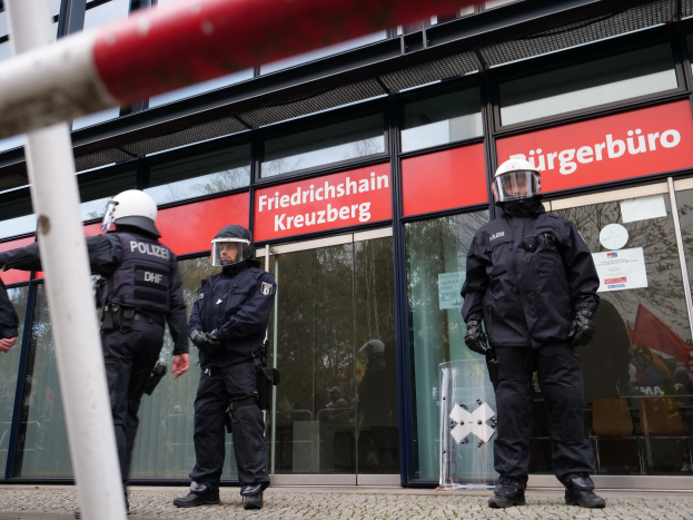 Polizeibeamte in Uniform stehen vor einem Glasgebäude, mit einem Pfahl links und Schildern mit der Aufschrift "Friedrichshain Kreuzberg" im Hintergrund.