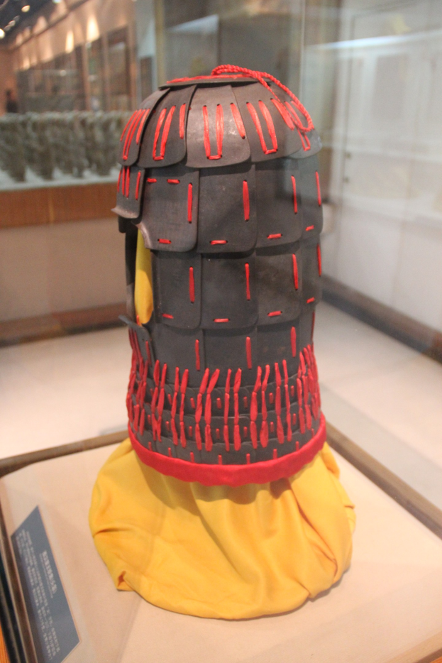 Helm in einer Glasvitrine mit einem gelben Tuch darauf präsentiert, begleitet von einer Texttafel auf der linken Seite und Deckenlampen im Hintergrund.