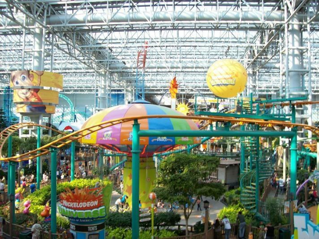 Ein großes Indoor-Wasserpark mit einer Achterbahn, umgeben von Menschen, Bäumen, Pflanzen und beleuchteten Deckenstangen und Lichtern.