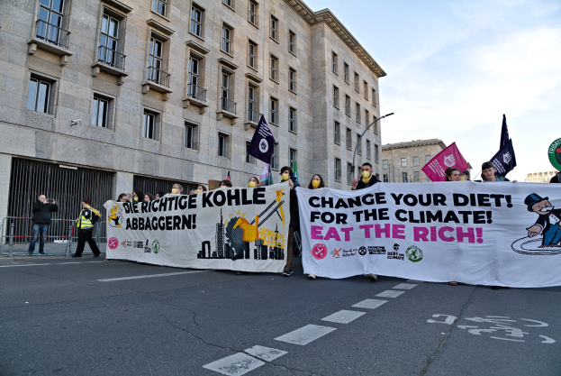 Gruppe von Menschen, die eine Straße entlanggehen und ein Banner mit der Aufschrift "Verändere deine Ernährung für das Klima, iss die Reichen" tragen, mit Gebäuden und Laternenmasten an der Straße und einem bewölkten Himmel.