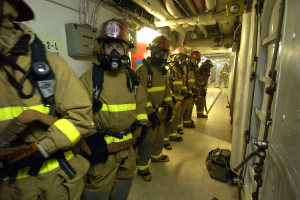 Eine Gruppe von Feuerwehrleuten in Helmen und Gasmasken geht einen Gang entlang mit einer Tür auf der rechten Seite, Taschen auf dem Boden und Rohren und Lampen im Hintergrund.