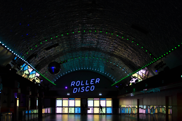 Ein Roller-Disco mit beleuchteter 'Roller Disco'-Anzeige, ein paar Menschen auf der Fläche, Glastüren im Hintergrund und Säulen links und rechts.