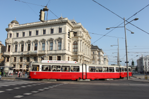 Eine rote und weiße Tram fährt auf einer von Gebäuden, Straßenlaternen und Lichtern gesäumten Stadtstraße, mit Kraftfahrzeugen, Fußgängern und Statuen unter einem klaren blauen Himmel.