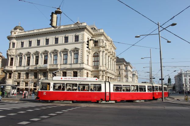 Eine rote und weiße Tram fährt auf einer von Gebäuden, Straßenlaternen und Lichtern gesäumten Stadtstraße, mit Kraftfahrzeugen, Fußgängern und Statuen unter einem klaren blauen Himmel.