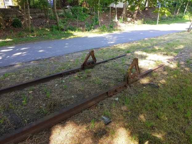 Bahnschiene auf einer Straße in Reparatur, mit Gras, Bäumen, einer Wand und einem Gebäude im Hintergrund.