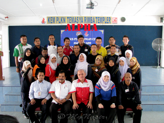 Eine Menschenmenge, die lachend vor einer Bühne posiert, mit einer Wandtafel mit Text, einem Tischventilator rechts, einem Podium links und einem Projektor oben.