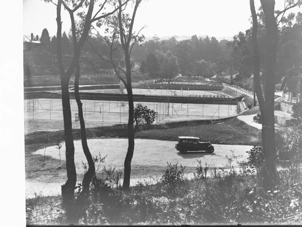 Ein Schwarz-Weiß-Foto eines vor einem Tennisplatz geparkten Autos, umgeben von Bäumen, Pflanzen, Gras, einem Zaun, Häusern und einem klaren Himmel.