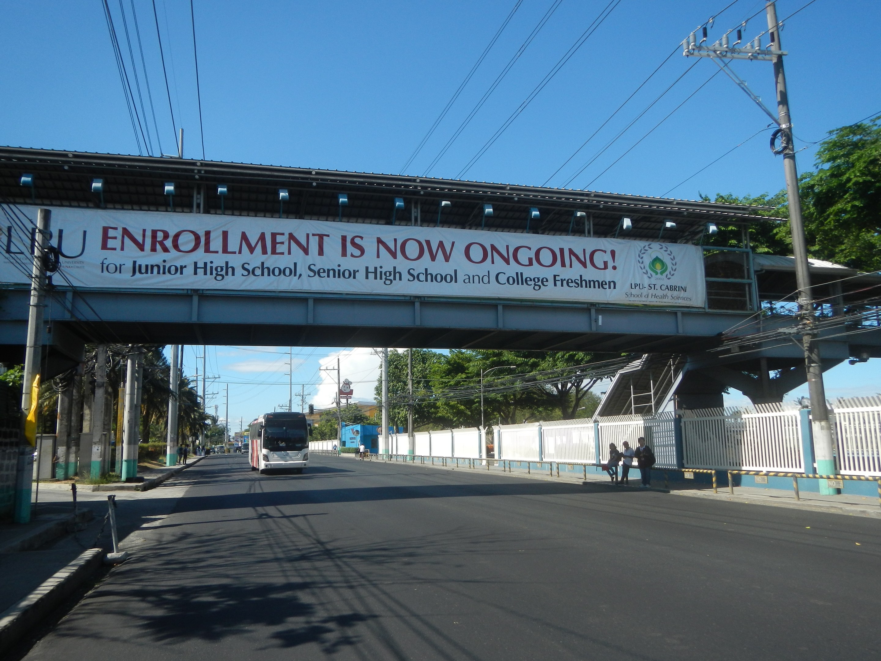 Ein Bus fährt unter einer Brücke mit einem Schild, auf dem "Jetzt Anmelden" steht, Menschen gehen auf einem Fußweg darunter, Strommasten mit Drähten, Bäume und ein klarer blauer Himmel im Hintergrund.