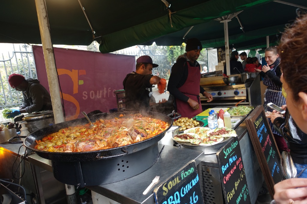 Ein Imbissstand mit einer Gruppe von Menschen, einer Person hält eine Flasche auf einer Kiste, ein Herd auf der rechten Seite, eine Tafel unten rechts und Essen unten links.