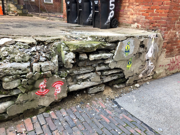 Ziegelwand mit Graffiti neben einem Ziegelgebäude, Straße im Vordergrund, schwarze Mülltonnen und Verkehrskegel im Hintergrund.