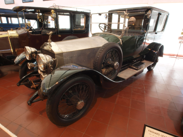 Ein Oldtimer mit glänzendem Lack und klassischem Design, der in einem Museum ausgestellt ist und von Texttafeln, einem Seil und gerahmten Ausstellungsstücken umgeben ist.
