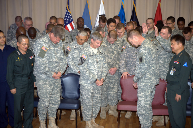 Eine Gruppe von Menschen in militärischer Kleidung steht zwischen roten und blauen Stühlen mit Flaggen im Hintergrund.