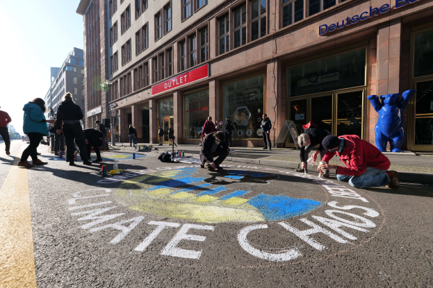 Eine Gruppe von Menschen sitzt vor einem Gebäude mit Fenstern und Namensschildern auf dem Boden, umgeben von Flaschen und anderen Gegenständen, mit Bäumen und einem klaren blauen Himmel im Hintergrund, während sie an einer Klimaprotest in Berlin teilnehmen.