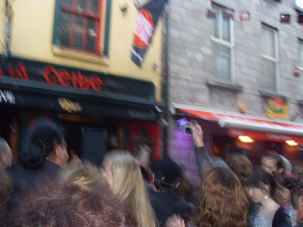 Ein unscharfer Bildausschnitt, der Menschen auf der Straße unten und Gebäude mit Lichtern, Fenstern und einer Flagge oben zeigt.