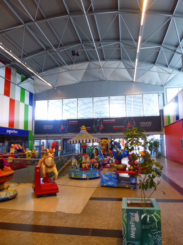 Großes Indoor-Vergnügungspark-Areal in einem Einkaufszentrum mit verschiedenen Attraktionen, einem Topf mit einer Pflanze, verstreuten Spielzeugen, Informationsschildern, Deckenleuchten und Metallstützen.