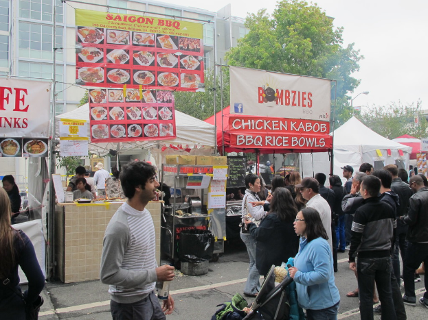 Menschen stehen auf einer Straße außerhalb der Stadt, mit einem Mann in einem grauen und weißen T-Shirt, der eine Wasserflasche hält und eine Frau in einer blauen Jacke, die einen Kinderwagen schiebt. Im Hintergrund sind Stände mit weißen und roten Zelten, farbige Banner und Gebäude, Bäume und eine Straßenlaterne zu sehen.