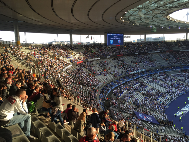 Große Menschenmenge in einem Stadion bei einem Fußballspiel, mit einer Bühne auf der rechten Seite, Fahnen, Stangen, einem Bildschirm und dem Allianz Arena in München, Deutschland im Hintergrund.
