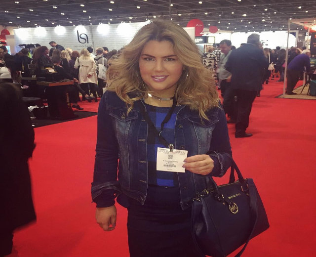 Frau auf einem roten Teppich an einer Modenschau, die lachend eine Handtasche hält, mit Menschen, Lichtern und Schildern im Hintergrund.