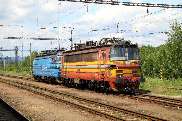 Ein Zugtriebwagen auf Schienen mit elektrischen Masten, Kabeln, B├Ąumen, Pflanzen und Wolken am Himmel.