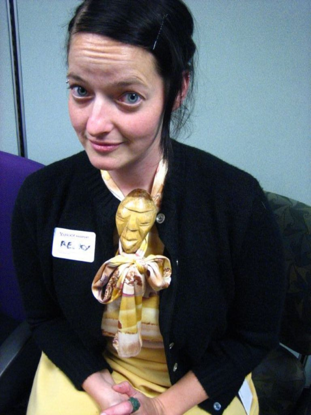 Eine Frau sitzt auf einem Stuhl und trägt eine schwarze Jacke und gelbes Kleid, mit einem Abzeichen an ihrer Kleidung, vor einer Wand.