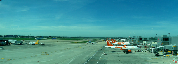 Eine Luftaufnahme eines Flughafens mit Flugzeugen, Gebäuden, Masten, Fahrzeugen und Bäumen, einem Himmel oben, einer Straße unten und einem Zaun im Vordergrund.