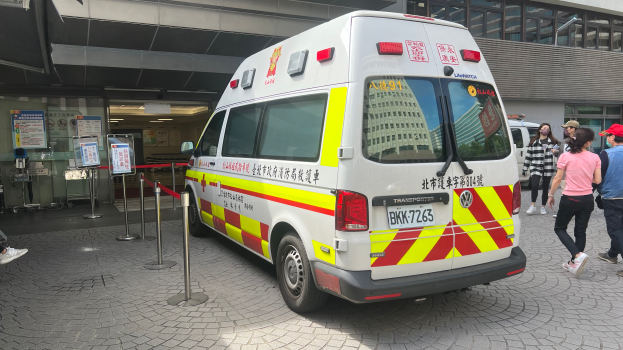 Eine Ambulanz steht vor einem Gebäude, mit Menschen auf der rechten Seite und einer Person auf einem Stuhl im Hintergrund; links an der Wand hängen Schilder mit Text und Postern, auf denen «Ambulanz-Service in Hong Kong» steht.