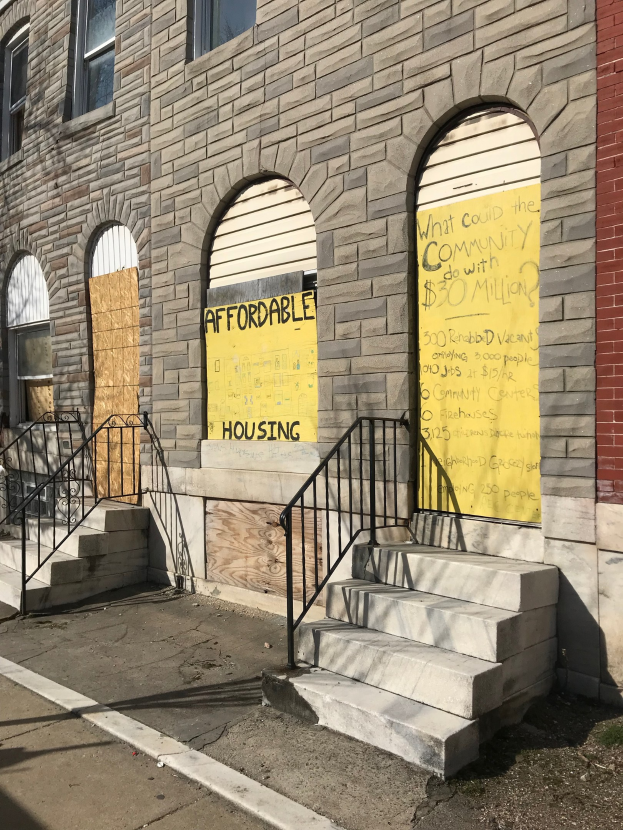 Ein verbarrikadiertes Gebäude mit einer Tafel, die "Bezahlbarer Wohnraum" anzeigt, eine Treppe zum Eingang und eine Straße unten.