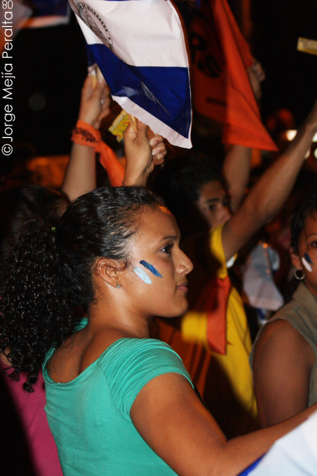 Eine Frau in einem gr├╝nen T-Shirt steht auf einer Demonstration und h├Ąlt ein wei├čes Schild in der Hand, mit vielen anderen Menschen hinter ihr.