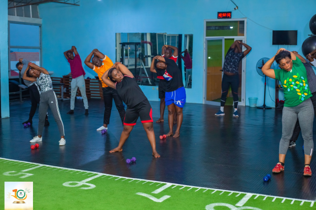 Gruppe von Menschen auf einem Gymnastikboden mit Hanteln während einer Yoga-Sitzung, umgeben von Gymnastikbällen, einem Ventilator, einem Fernseher, einer Tür, einem Spiegel und Deckenleuchten.