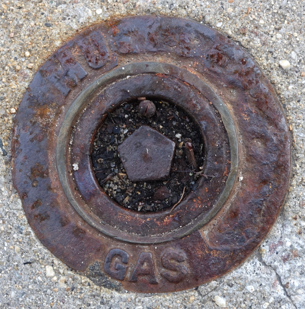 Ein verrosteter Metall-Schachtobdeckel auf dem Boden mit dem Wort 'Gas' darauf.