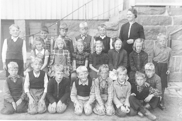 Ein Schwarz-Weiß-Foto einer Gruppe von Kindern, die vor einem Gebäude mit einer Wand und Treppen mit Geländern posieren, mit dem Text "1930er Schulkinder" unten.