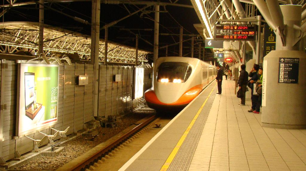 Mehrere Personen stehen auf einem Bahnsteig mit einem Zug auf den Schienen; links hat eine Wand ein Schild, rechts sind einige Schilder an Pfählen befestigt.