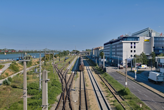 Ein Zug fährt auf Schienen neben einer Stadtlandschaft mit Gebäuden, Straßeninfrastruktur, Fahrzeugen, Grünflächen, Beschilderung, einer Brücke und einem bewölkten Himmel.