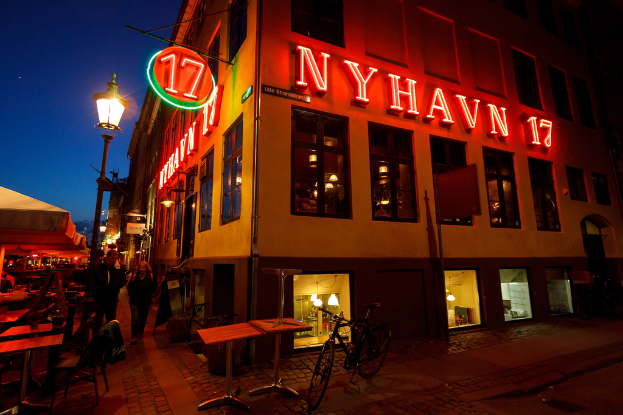 Ein Gebäude mit rotem Text an der Fassade, begleitet von einem Fahrrad, Tischen und Stühlen, mit einer Straßenansicht daneben.