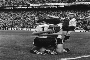 Drei Fußballspieler feiern ein Tor auf dem Boden, umgeben von einer jubelnden Menge und schwenkenden Fahnen im Hintergrund, dargestellt in Schwarz-Weiß.