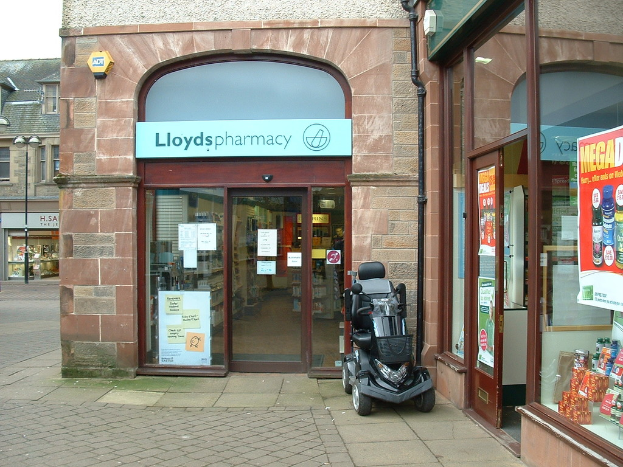 Eine Apothekenfiliale mit einem Fahrzeug davor und einem Gebäude in der linken Ecke.