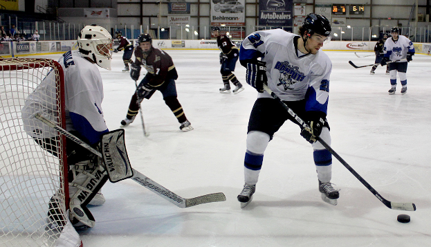 Eine Gruppe junger Männer, die auf einem Eisplatz Eishockey spielt, trägt Helme und hält Hockey-Schläger, links steht ein Torpfosten und im Hintergrund hängen Banner.