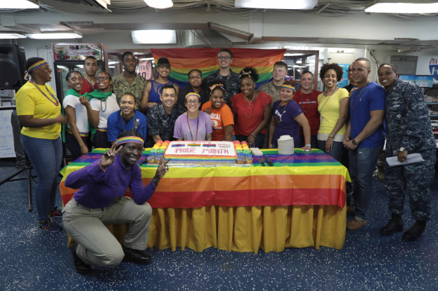 Eine Gruppe von Menschen, die vor einem Tisch mit einem "Pride Month"-Kuchen, Bannern und Lichtern posieren.