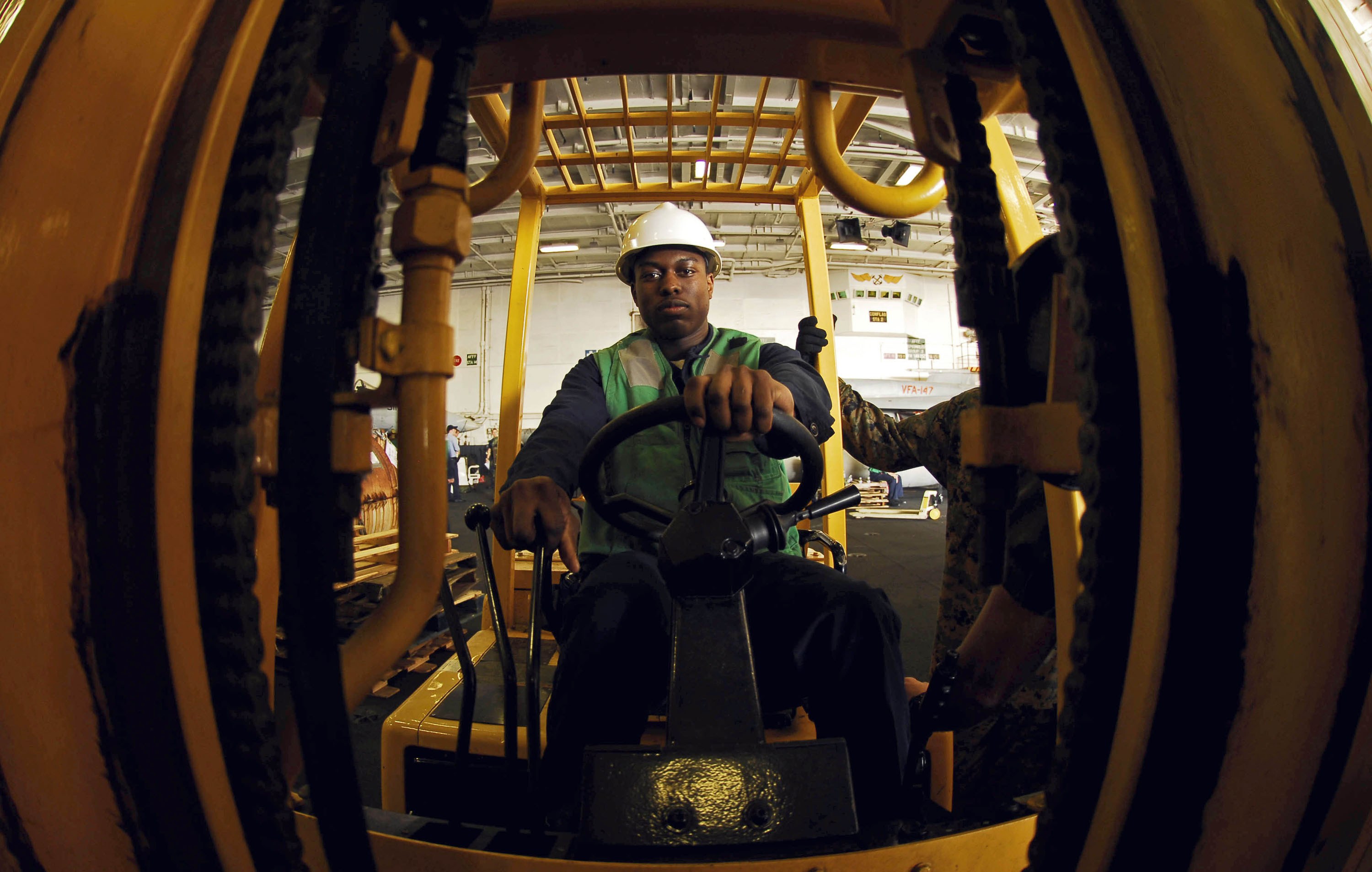 Ein Mann in einer grünen Jacke und einem weißen Helm bedient einen Gabelstapler in einer industriellen Umgebung mit anderen Menschen im Hintergrund.