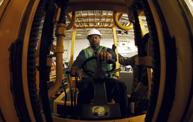 Ein Mann in einer grünen Jacke und einem weißen Helm bedient einen Gabelstapler in einer industriellen Umgebung mit anderen Menschen im Hintergrund.