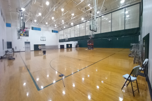 Große Turnhalle mit Holzboden, Stühlen, einer Wand, Deckenleuchten und einem Basketballfeld.