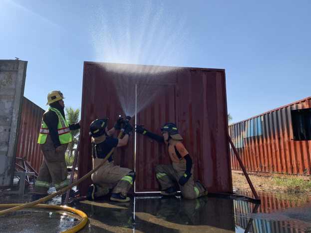 Feuerwehrleute in Helmen und Handschuhen spritzen Wasser aus einem Schlauch auf einen Container, um ein Feuer zu löschen, mit Bäumen und einem klaren blauen Himmel im Hintergrund, einem Rohr auf der linken Seite und einem Metallcontainer auf der rechten Seite.