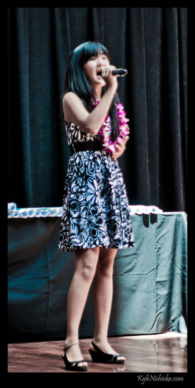 Eine Frau in einem bunten Kleid singt auf der B├╝hne in ein Mikrofon, mit einem Tisch und Papieren im Hintergrund hinter einem Vorhang.