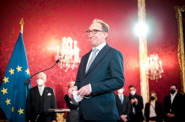 Ein Mann im Anzug und Krawatte steht an einem Rednerpult, hält ein Blatt Papier in der Hand und hat links ein Mikrofon, während er einer maskierten Zuhörerschaft in einem Raum mit Kronleuchtern eine Rede hält.