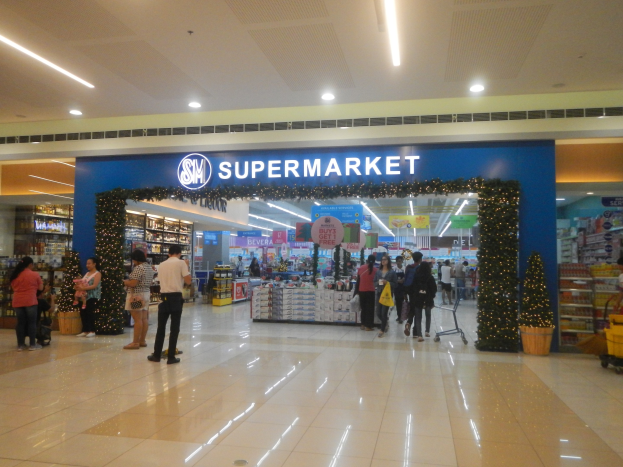 Menschen vor einem Supermarkt mit Taschen, einige halten Tüten, mit Einkaufswagen, Topfpflanze, Regalen, Schildern und Deckenbeleuchtung im Hintergrund.