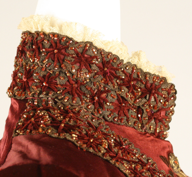 Ein Nahaufnahme eines roten und goldenen Kleides auf einer Schaufensterpuppe, das mit feiner Spitzenverzierung gegen einen weißen Hintergrund präsentiert wird.