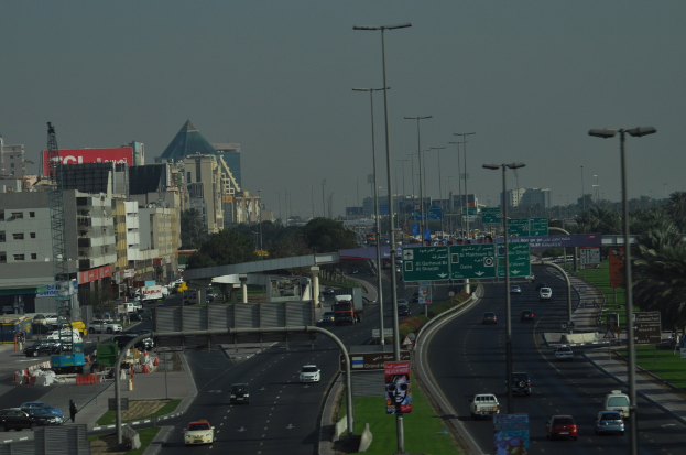 Eine Luftaufnahme einer Stadt mit Gebäuden, Türmen, Fahrzeugen, Werbetafeln, Schildern, Straßen, Bäumen, Straßenlaternen, Menschen, Verkehrsleitkegeln und Informationsanzeigen unter einem klaren Himmel.