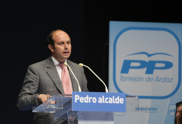 Ein Mann im Anzug spricht an einem Pult mit Mäusen und einem Glas darauf, mit schwarzem Hintergrund und einer Werbetafel rechts.