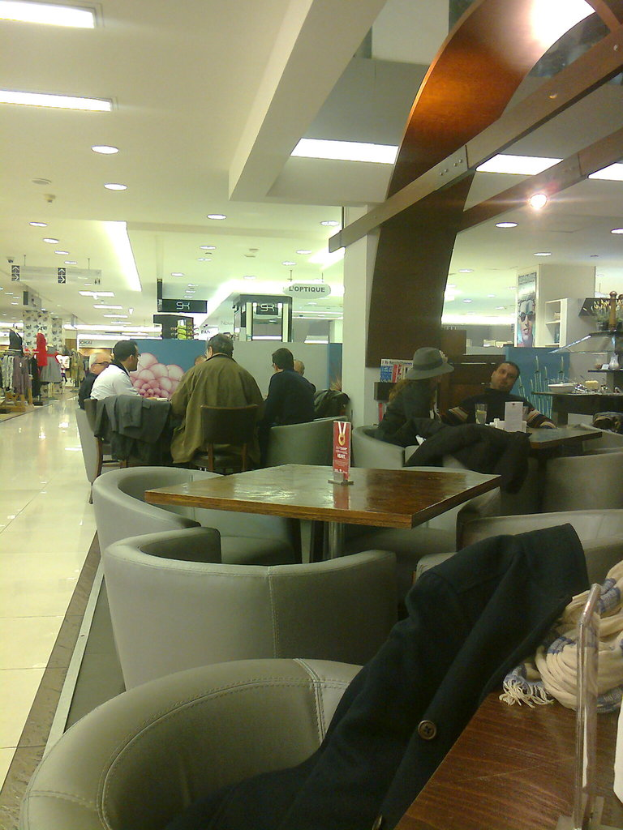 Leute sitzen auf Stühlen in einem Einkaufszentrum mit Kleidung auf einem Tisch, Geschäfte auf der linken Seite und Lampen an der Decke.