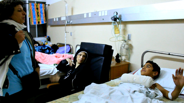 Ein Krankenhauszimmer mit mehreren Betten, Infusionsflaschen auf Ständern zwischen den Betten und einer Frau mit einer Tasche in der Nähe eines Patienten.